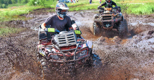 Two people wearing helmets and goggles, riding ATVs through large puddles in a muddy off-road course.