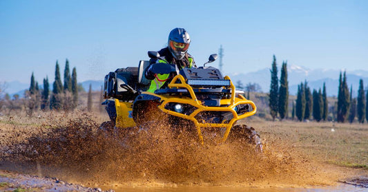 A woman wearing a helmet and protective gear drives a bright yellow ATV through the mud. The ATV's radiator is raised.