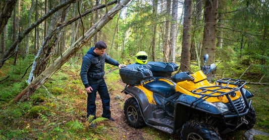 A yellow ATV is stopped on a trail in the woods. The rider stands with his hand on the back of the ATV as he examines it.