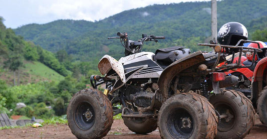 Two dirt-covered, lifted ATVs standing on packed dirt ground. They face a valley with tree-covered hills rising in the distance.